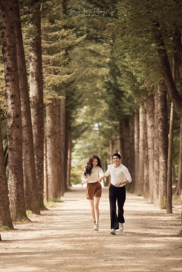 two people walking down a dirt road between tall tree's and one person is holding the hand of another