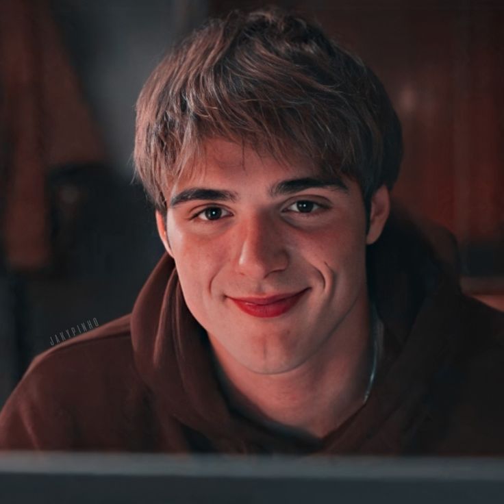 a young man wearing a brown hoodie looks at the camera while sitting in front of a computer screen