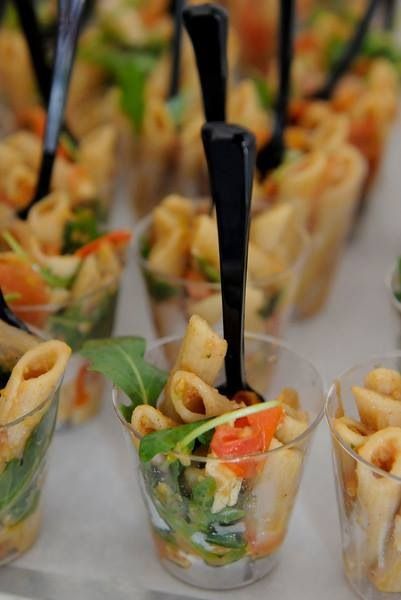 small cups filled with pasta and vegetables on top of a white table covered in black plastic utensils