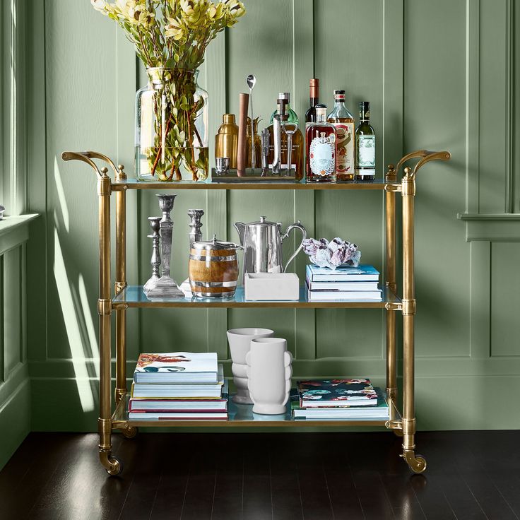 a bar cart filled with bottles and glasses on top of a hard wood floor next to a green wall