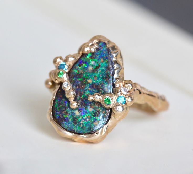 "Our one-of-a-kind 14k yellow gold boulder-opal ring celebrates the natural beauty of this incredible gemstone with a gracefully undulating branch band and hand-hammered texture that emphasizes it's slightly rustic nature: organic minimalism with a brutalist vibe. It's designed to stand on its own as a unique statement, but it also plays well with other pieces adding texture to your daily ring stack. The companion stones, diamond, blue topaz and tsavorite garnet, play to the galactic color of th Organic Minimalism, Boulder Opal Ring, Mode Kimono, Branch Ring, Tsavorite Garnet, Organic Style, Ring Stack, Party Rings, Raw Beauty