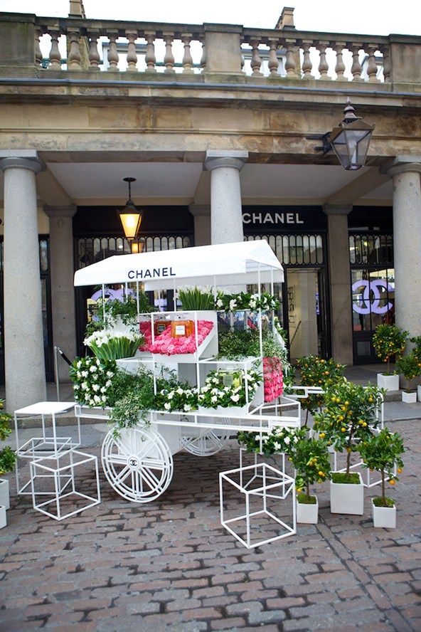 a white cart filled with flowers sitting on top of a brick road next to a building