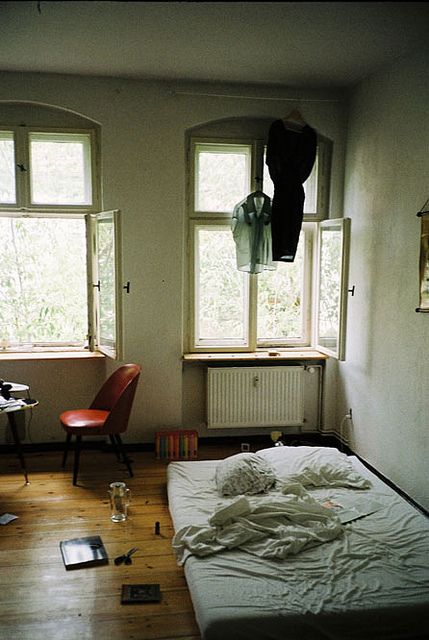 an unmade bed sitting next to two windows in a room with hard wood floors