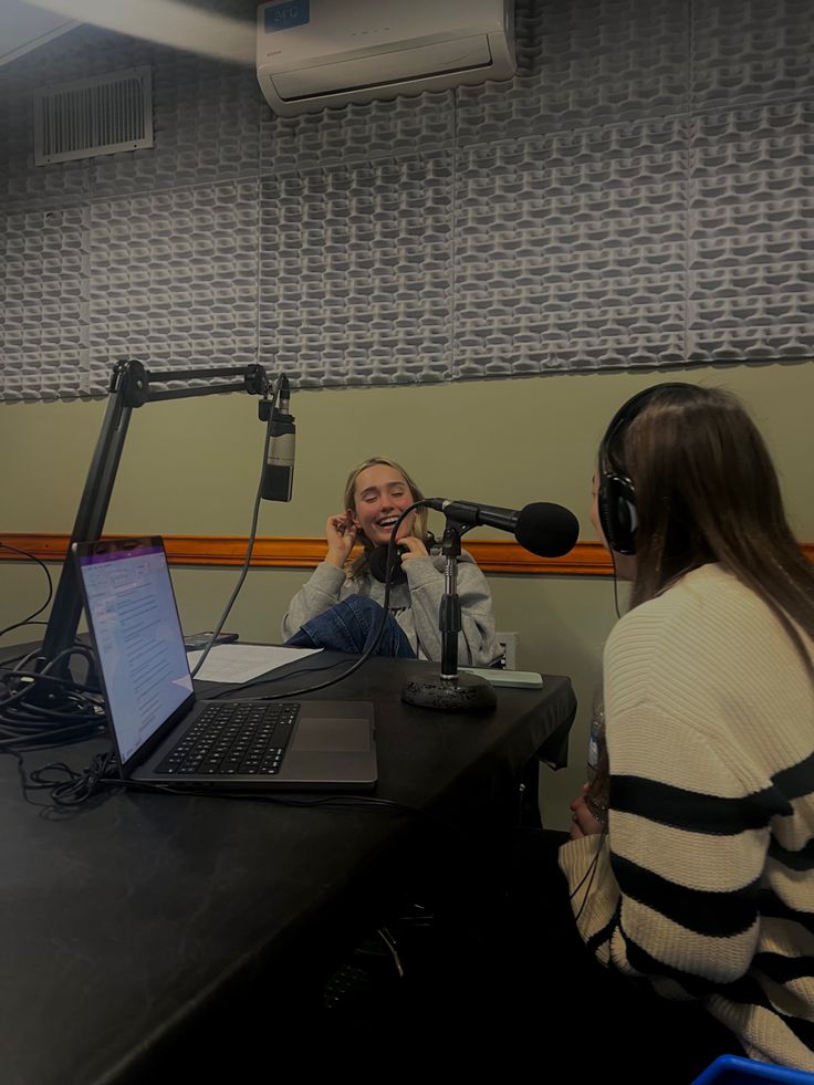 two women sitting at a table in front of microphones talking to each other and laughing