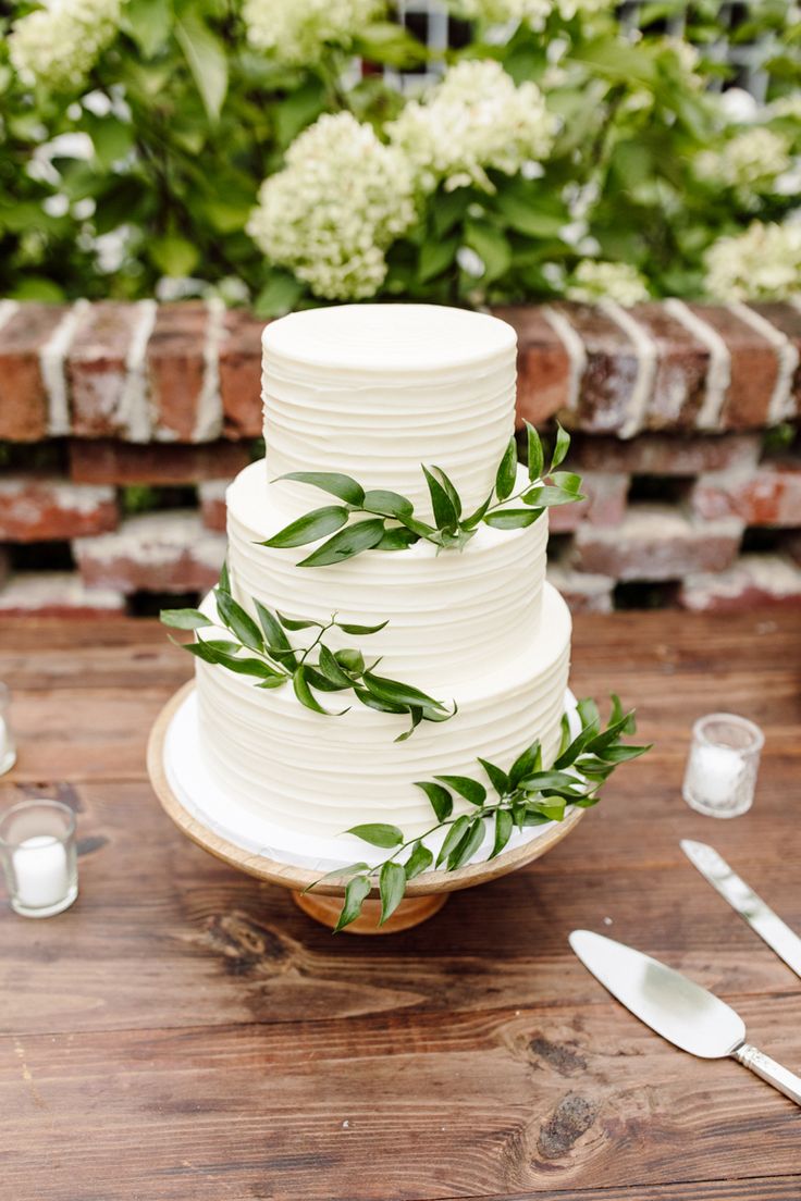 a white wedding cake with greenery on top sits on a table next to silverware