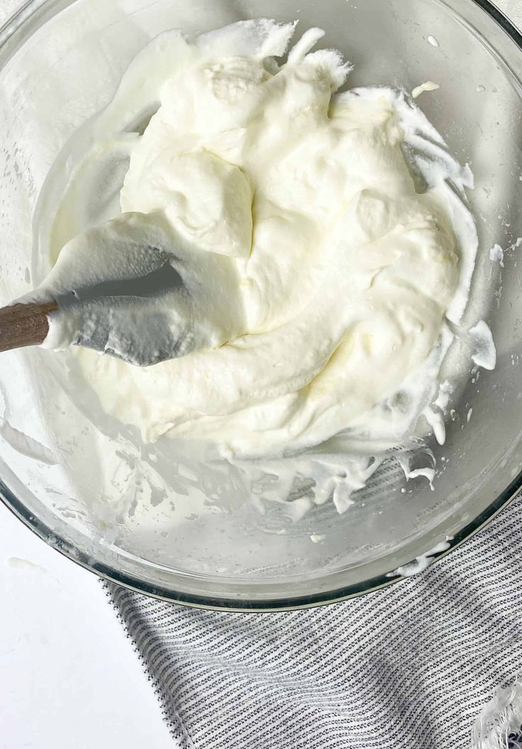 a glass bowl filled with whipped cream on top of a gray towel next to a wooden spoon