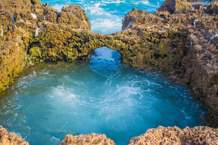 an aerial view of the ocean and rocks with water coming out from under that bridge