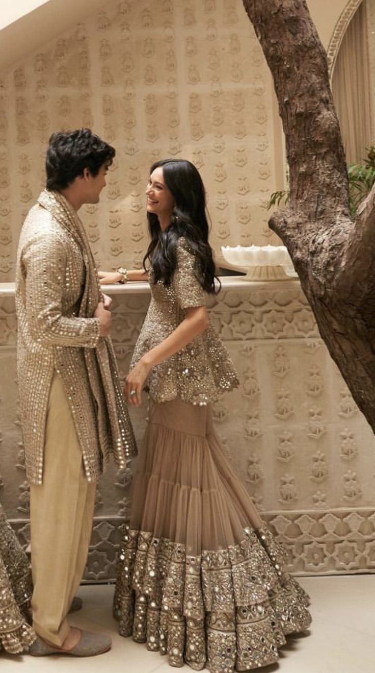 two people standing next to each other in front of a tree and wall with gold sequins on it