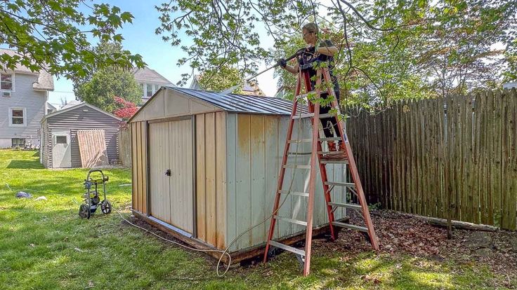 a man standing on a ladder next to a shed in the grass near a fence