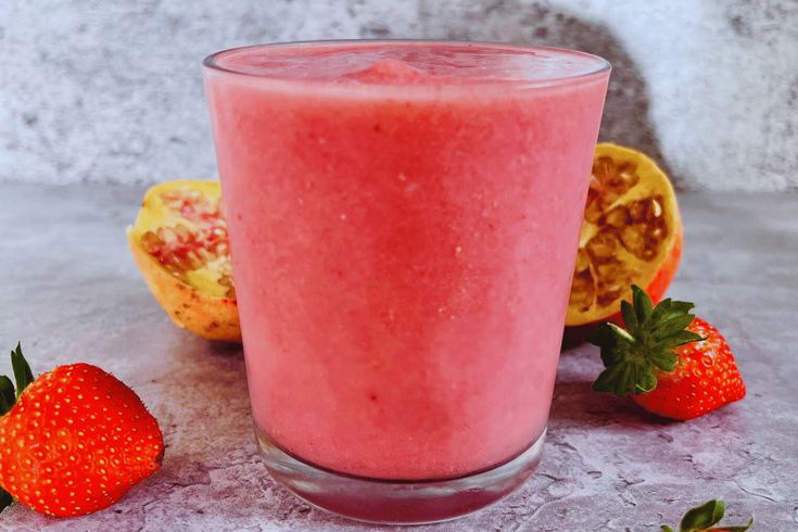 a pink smoothie in a glass next to some strawberries and a pomegranate