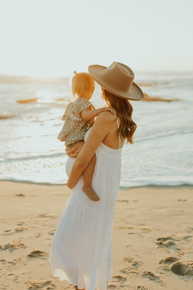 a woman in a white dress and hat holding a baby on the beach at sunset