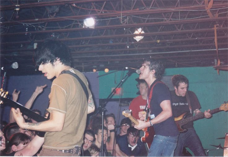 two young men playing guitars in front of an audience at a music concert, while one man is singing into a microphone