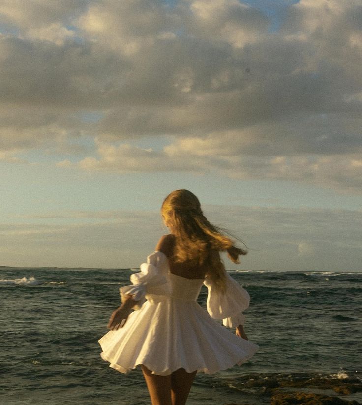 a woman in a white dress standing on the beach looking out at the ocean with her back to the camera