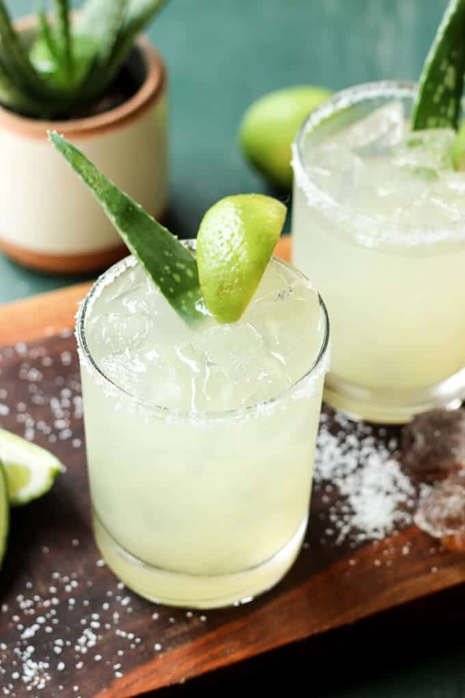 two margaritas are garnished with an aloey and lime slice on a cutting board