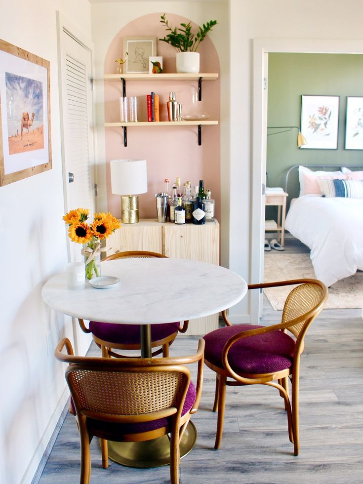 the dining table is next to two chairs and a shelf with books on it, in front of a bedroom