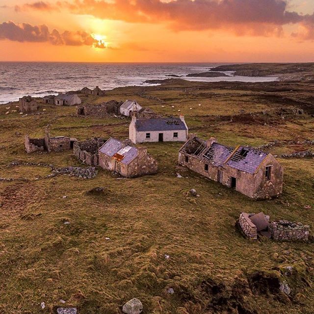 the sun is setting over an old farm by the ocean with abandoned buildings in the foreground