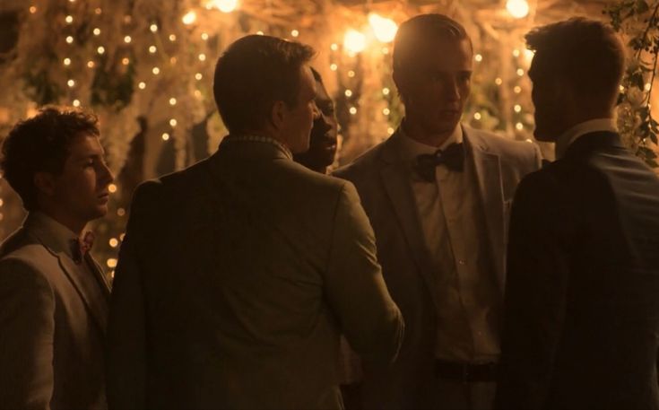 three men in tuxedos are talking to each other at a dinner table with lights on the wall behind them