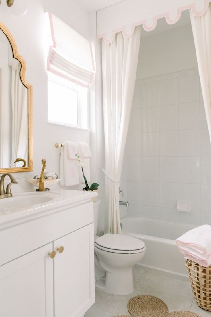 a white bathroom with pink accents and gold accessories on the sink, toilet, tub, and shower curtain