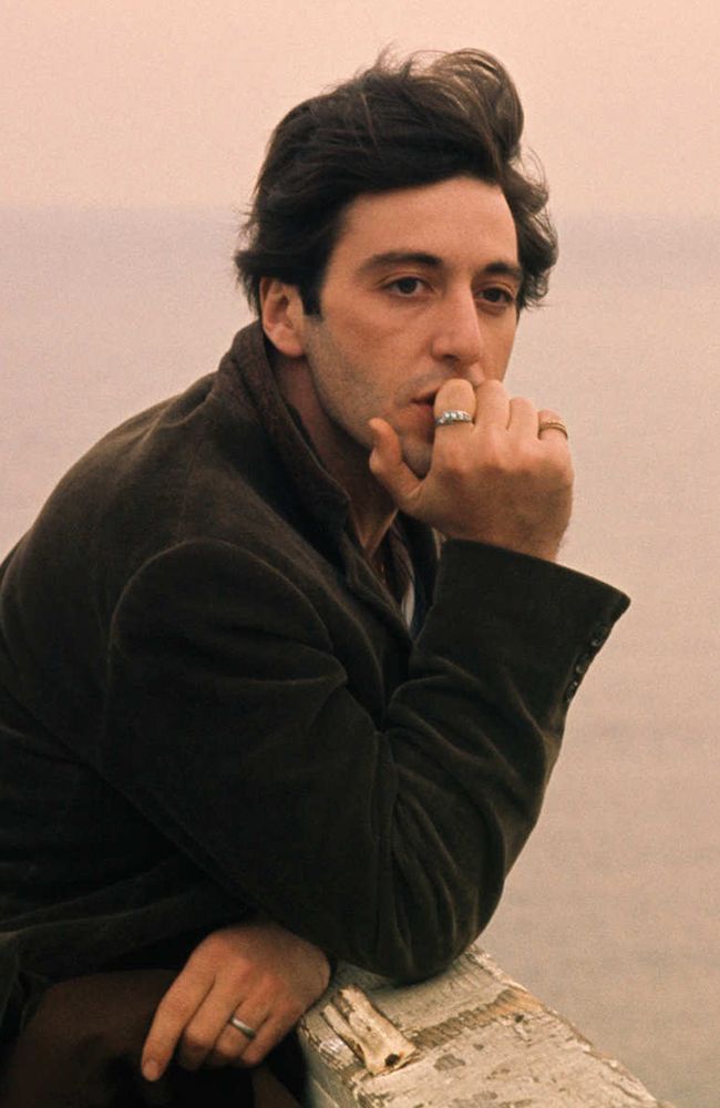 a man sitting on top of a wooden bench next to the ocean with his finger in his mouth