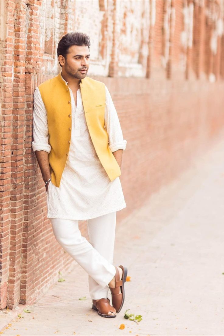 a man leaning against a brick wall wearing a yellow vest and white pants with brown shoes