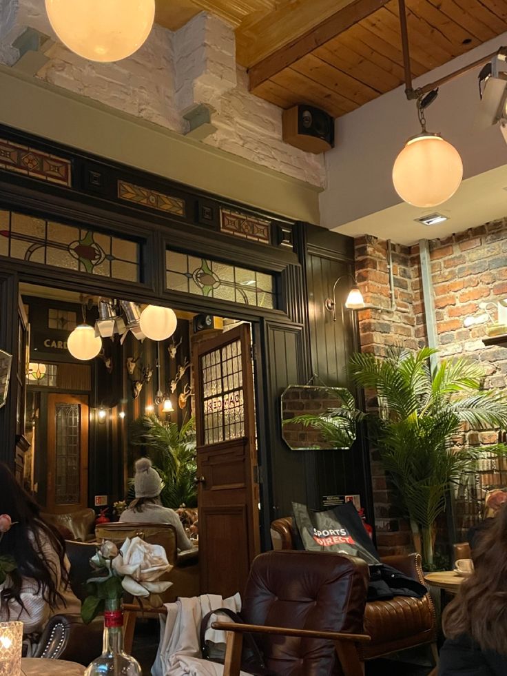 people sitting at tables in a restaurant with lights hanging from the ceiling and windows above them