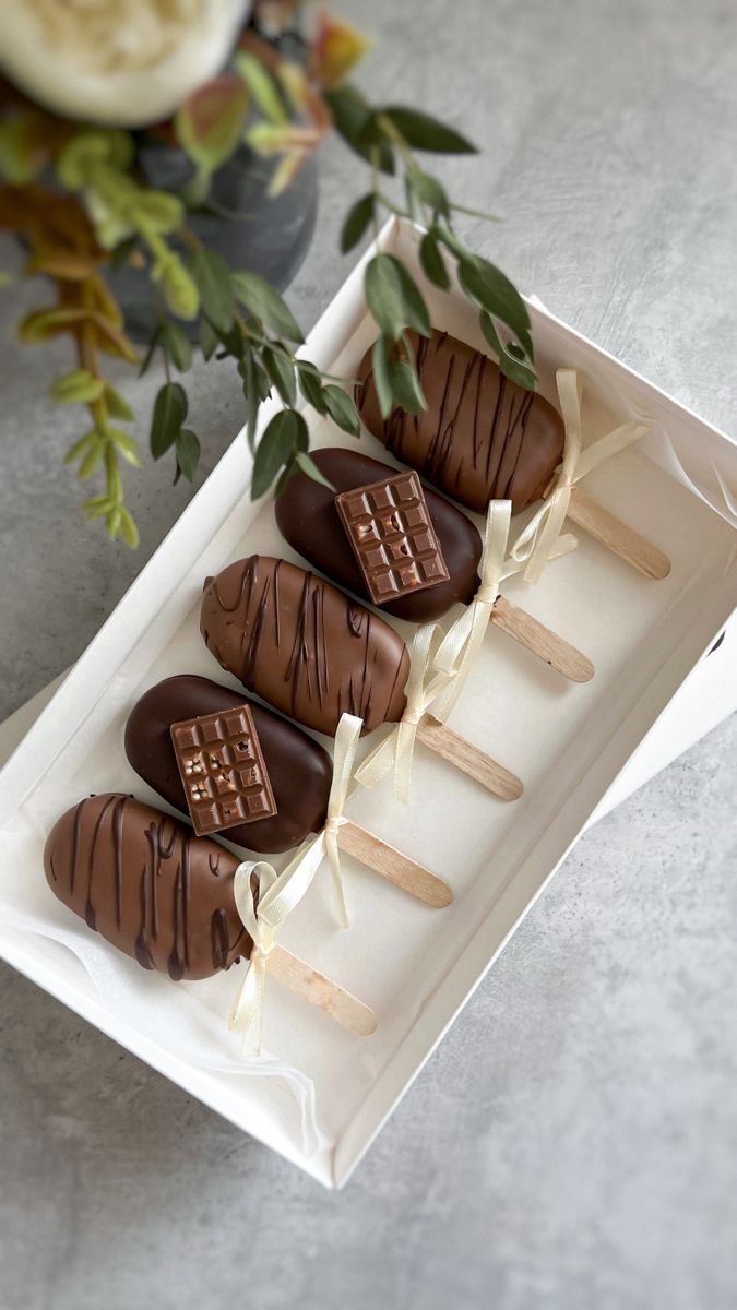 four chocolate covered desserts on sticks in a white box next to flowers and greenery