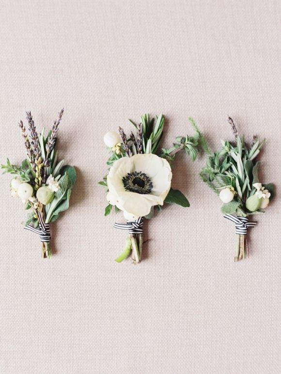 three boutonnieres with white flowers and greenery are arranged on a beige surface