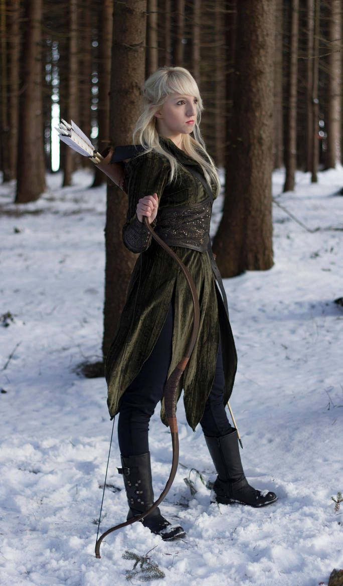 a woman dressed in green is holding a bow and arrow while standing in the snow