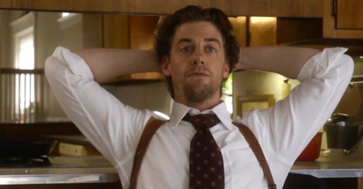 a man with suspenders and a tie is holding his hands behind his head in the kitchen