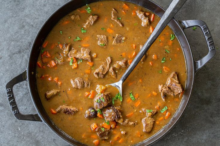 a pot filled with meat and carrots on top of a table next to a spoon