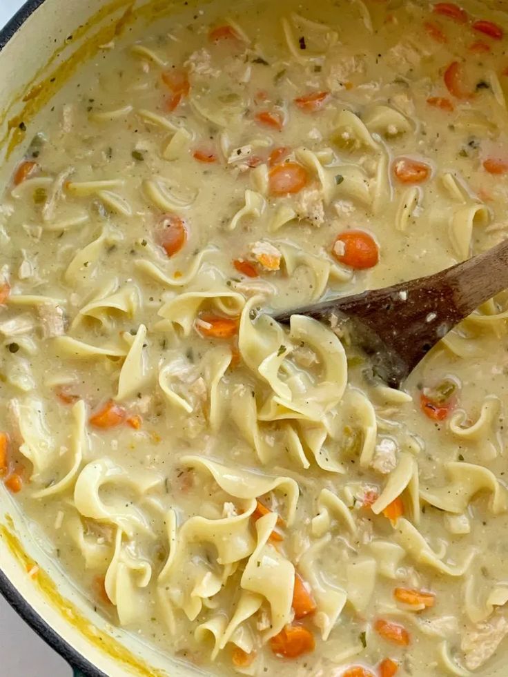 chicken noodle soup in a pot with a wooden spoon on the side and carrots