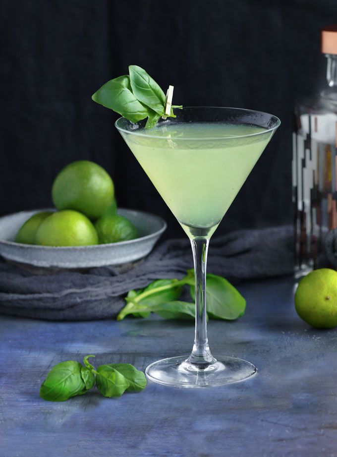 a green drink in a martini glass with basil on the rim and limes next to it