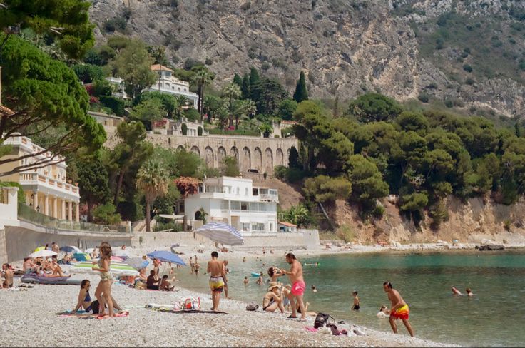 many people are on the beach and in the water near some buildings, trees, and mountains