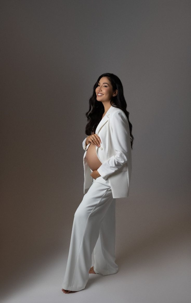 a pregnant woman posing for a photo in white pants and blazer with her hands on her hips