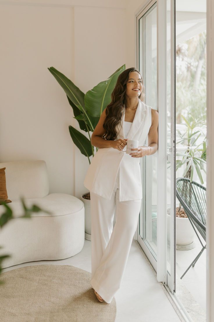 a woman standing in front of a glass door wearing white pants and a top with one hand on her hip