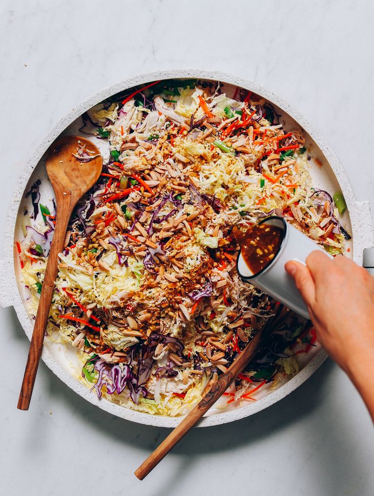 two hands holding wooden spoons over a large bowl of shredded cabbage and carrots