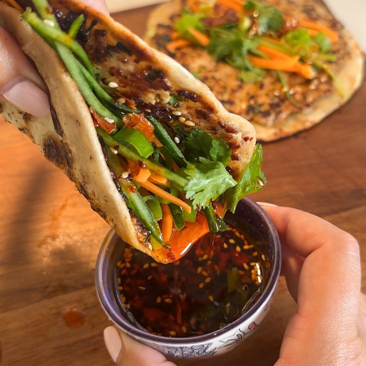 a hand holding a small bowl filled with food next to two flatbreads on top of a wooden table