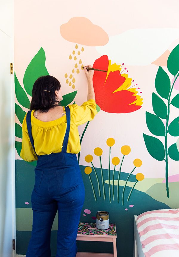 a woman in overalls painting a flower on a wall with flowers and plants painted on it