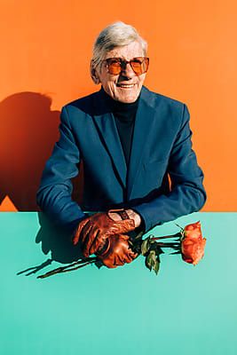 an older man wearing gloves and holding a rose in front of a green table with orange walls