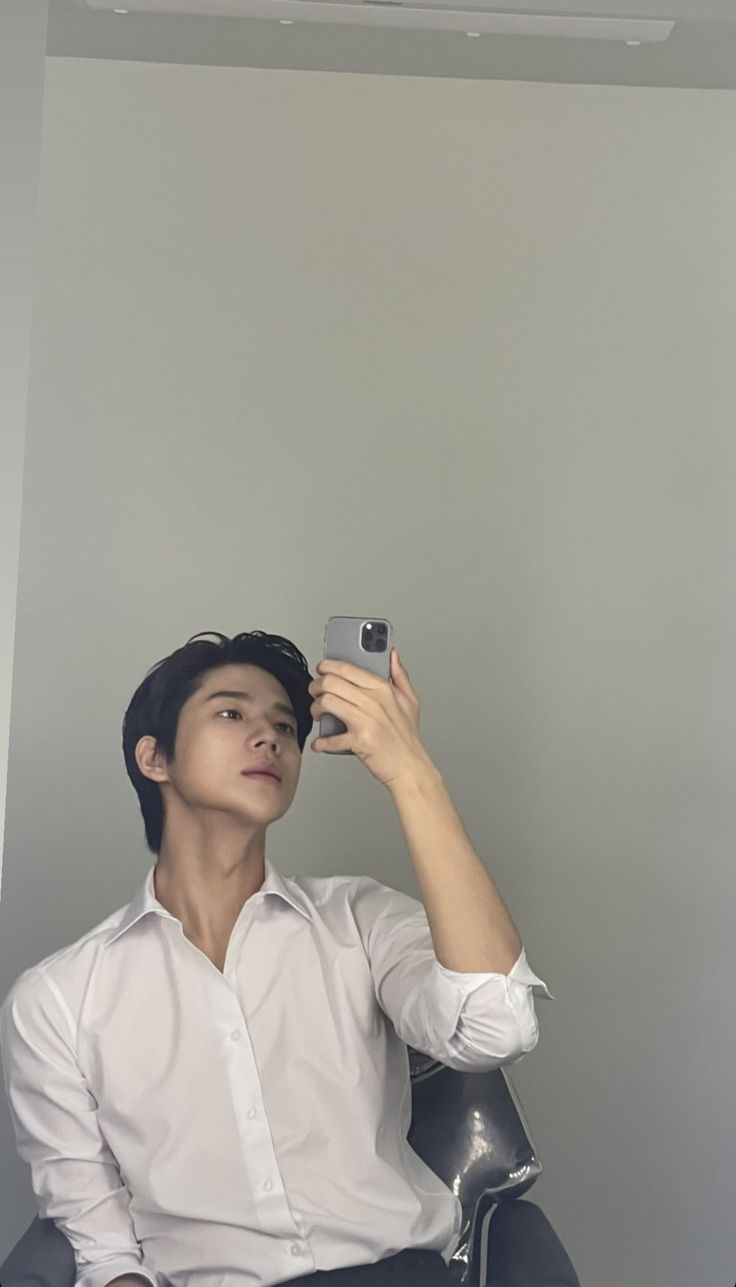 a man sitting in a chair taking a selfie with his cell phone while wearing a white shirt and black pants