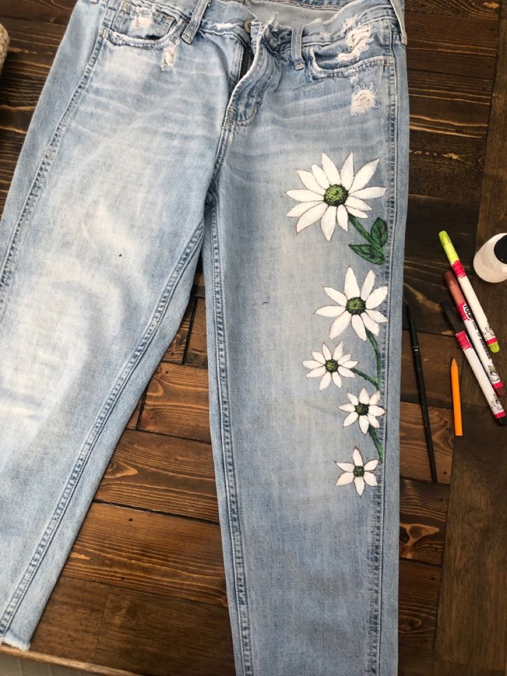 a pair of jeans with white daisies painted on them next to markers and pencils