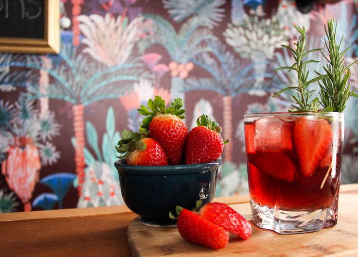 strawberries in a bowl next to a glass filled with liquid and rosemary sprigs