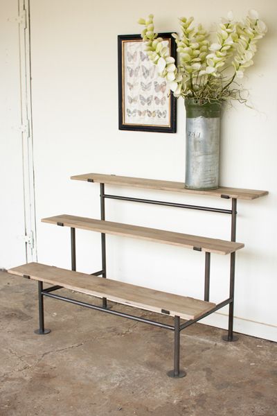 a metal vase with flowers on top of a wooden shelf next to a wall mounted art piece