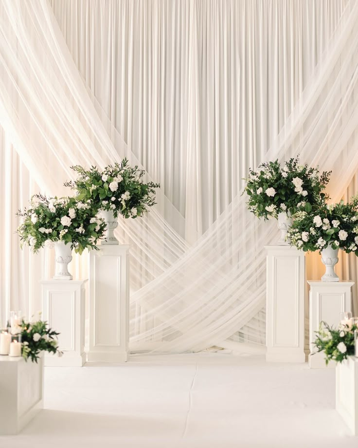 two white vases with flowers and greenery on them in front of a curtain