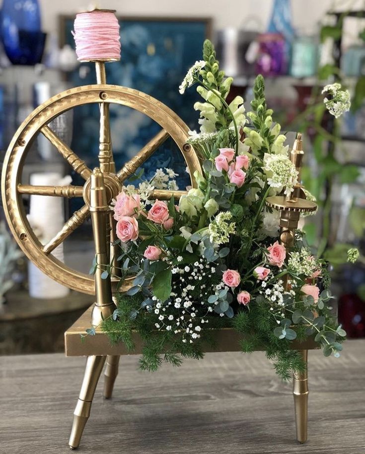 an arrangement of flowers and greenery on a table with a spinning wheel in the background