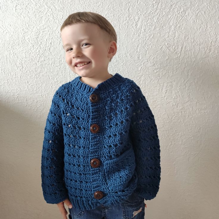 a young boy wearing a blue crocheted cardigan and jeans standing in front of a white wall