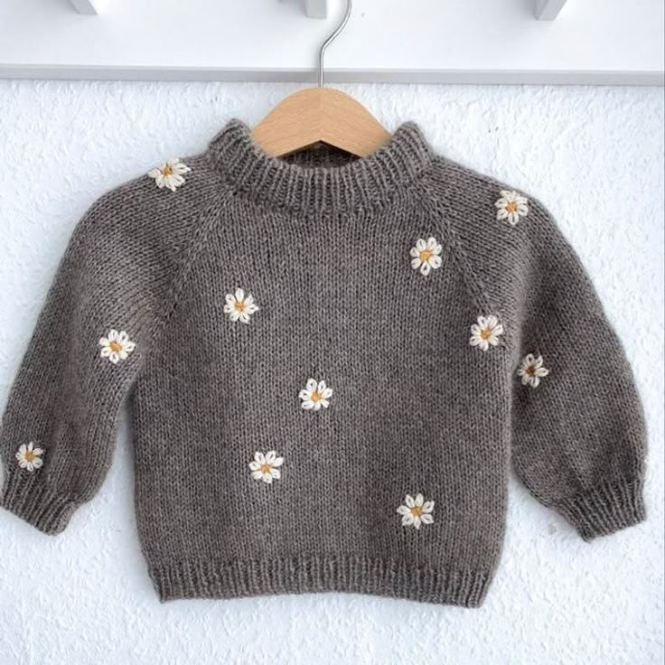 a gray sweater with daisies is hanging on a white coat hanger next to a wall