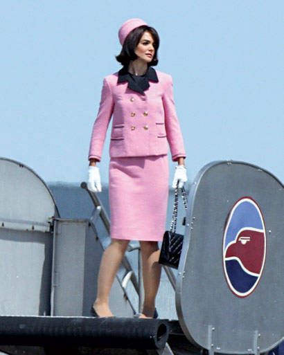 a woman in a pink suit and hat standing on top of a metal structure with an american flag behind her