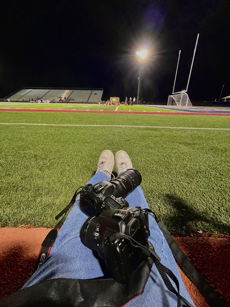 a person sitting on top of a baseball field with a camera in their lap and the lights behind them