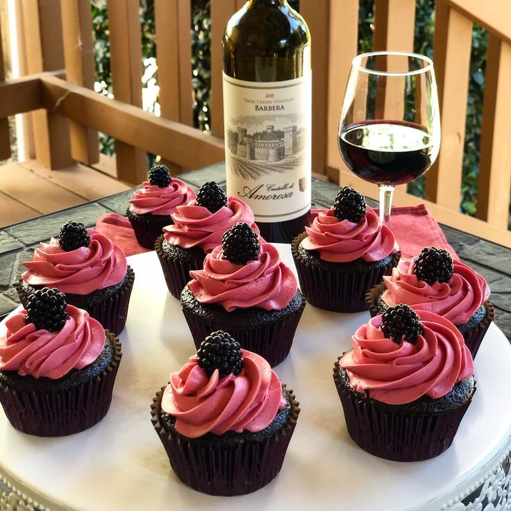 cupcakes with pink icing and blackberries are on a plate next to a bottle of wine